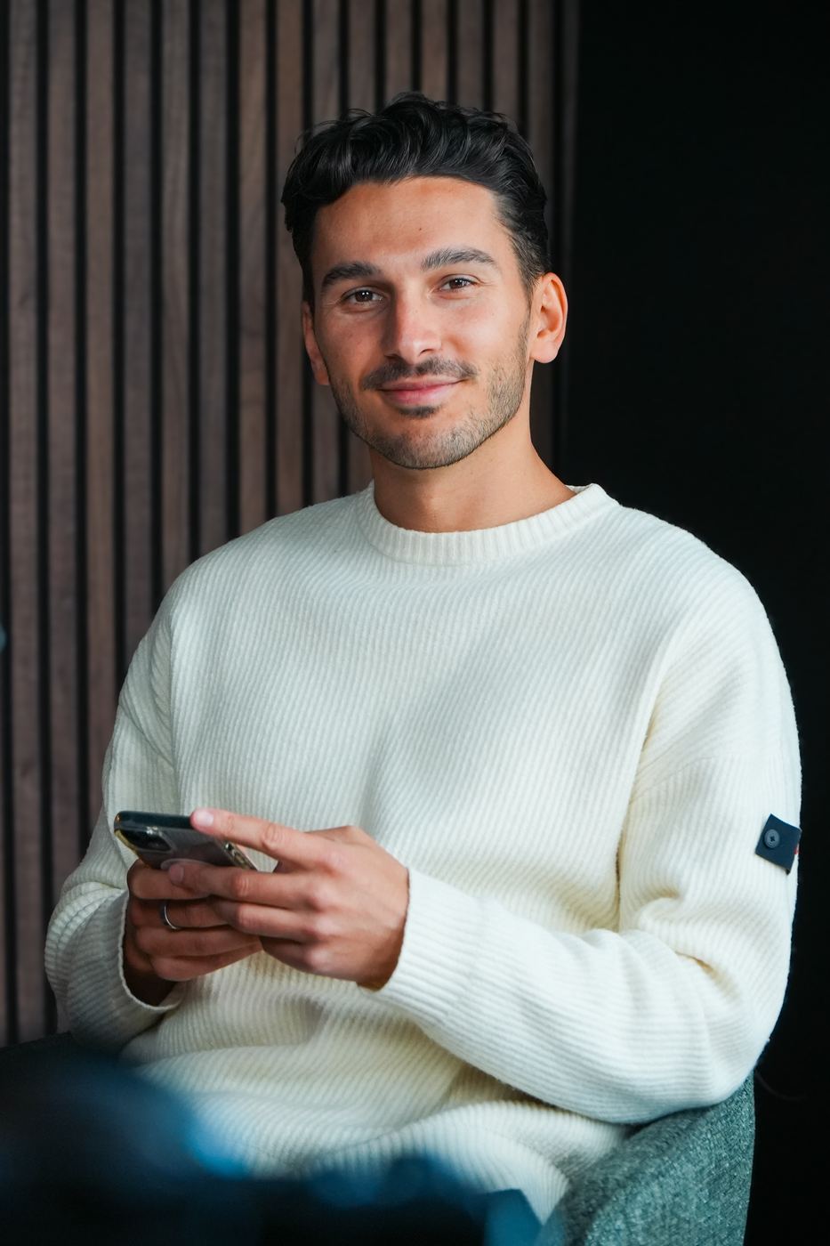 Frederic Luhede im hellen Strickpullover, lächelnd, mit Smartphone in der Hand